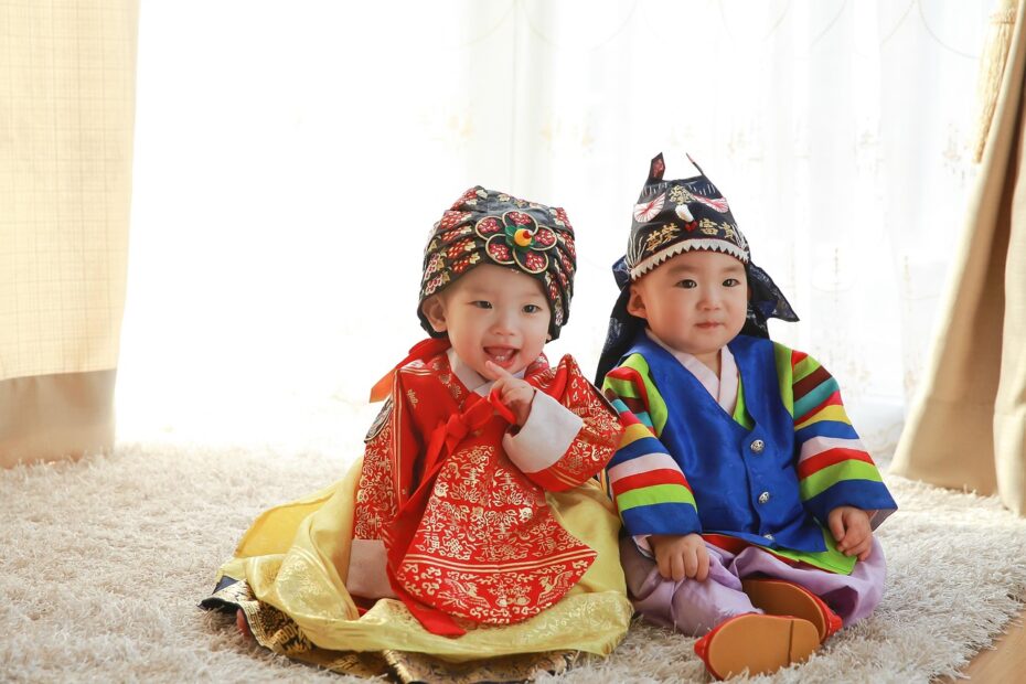 붉은 한복과 색동저고리 한복을 입고 전통 모자를 쓴 아기 두 명이 카펫 위에 앉아 있는 모습